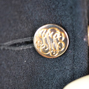 May include: A gold-colored button with a monogram design on a black fabric background.
