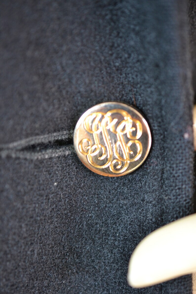 May include: A gold-colored button with a monogram design on a black fabric background.