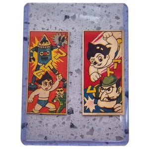 May include: Two vintage Japanese Astro Boy trading cards. The cards feature colorful illustrations of Astro Boy and other characters in action poses, set against a red background. The cards are encased in a clear plastic holder.