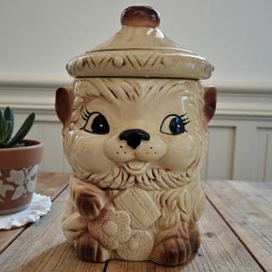 May include: A vintage ceramic cookie jar shaped like a smiling cartoon animal with large eyes and a black nose. The jar is beige with brown accents and a matching lid. The animal is holding a cookie.