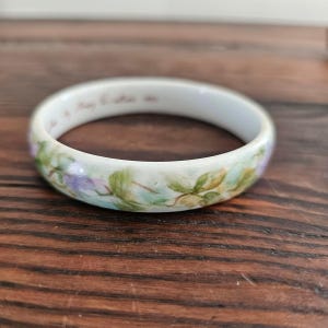 May include: A white ceramic bangle bracelet featuring a hand-painted floral pattern with green leaves and purple accents. The interior of the bracelet displays faint, cursive text written in a light brown ink along the inner rim of the band.