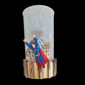 May include: A clear, cylindrical container with a Superman figurine in flight. The figure is in a blue suit with a red cape, set against a gold-colored geometric globe. The base has a decorative, scalloped design.