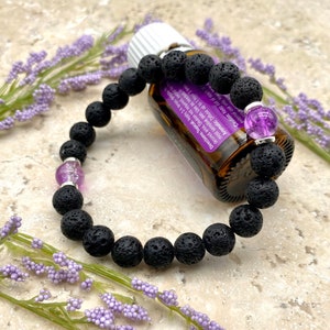 May include: Black lava rock bracelet with two amethyst beads and a silver charm. The bracelet is lying on a table next to a small bottle of essential oil.