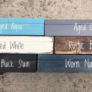 May include: A collection of stained wood samples in various colors, including Aged Aqua, Aged Grey, Aged White, Rustic Brown, True Black Stain, and Worn Navy. Each sample has the color name written in white script.