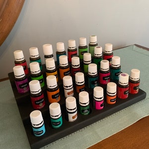 May include: A tiered, dark-stained wooden essential oil display stand holding numerous small glass bottles with white caps and colorful labels. The bottles are arranged in rows, showcasing a variety of essential oil blends.