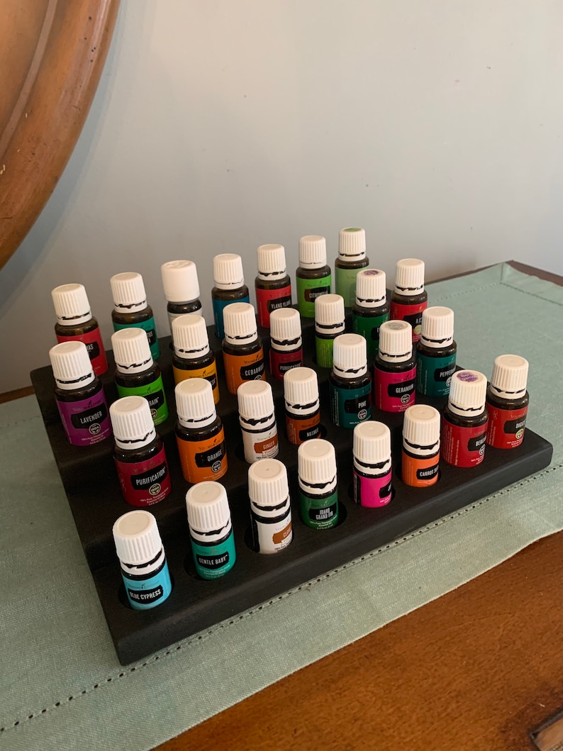 May include: A tiered, dark-stained wooden essential oil display stand holding numerous small glass bottles with white caps and colorful labels. The bottles are arranged in rows, showcasing a variety of essential oil blends.