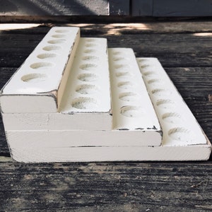May include: A tiered, white wooden display stand with multiple circular cutouts. The stand has a distressed finish, with visible wear and tear. The design features three levels, each with a series of evenly spaced holes, likely for holding small items.