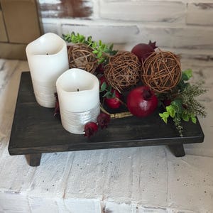May include: A decorative wooden tray with two white pillar candles, decorative brown woven spheres, red pomegranates, and green foliage. The tray is dark gray and sits on a white brick surface.