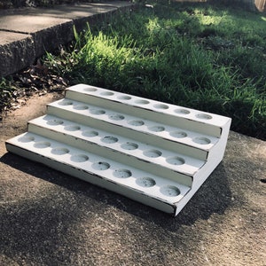 May include: A white, tiered wooden display stand with distressed edges. The stand has three levels, each with multiple circular holes. The stand is designed to hold small items and is placed on a concrete surface.