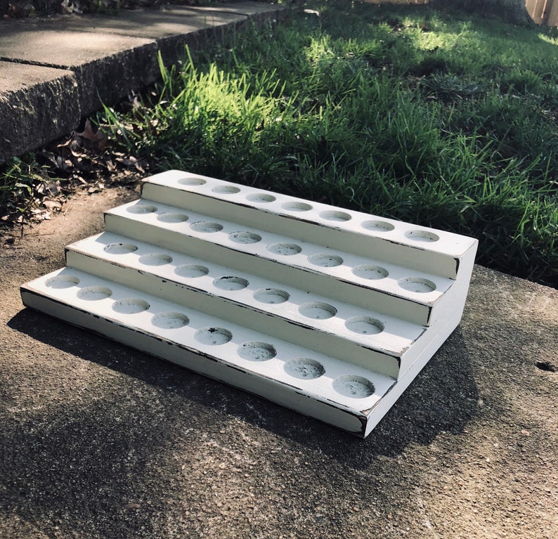 May include: A white, tiered wooden display stand with distressed edges. The stand has three levels, each with multiple circular holes. The stand is designed to hold small items and is placed on a concrete surface.