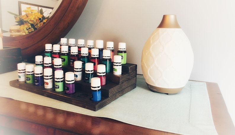 May include: A wooden tiered display holds numerous small essential oil bottles with white caps and colorful labels. A white and gold essential oil diffuser sits to the right. The items are arranged on a wooden surface.