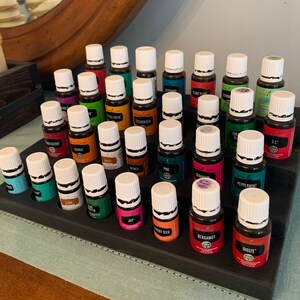 May include: A tiered, dark-stained wooden essential oil display stand holding numerous small glass bottles with white caps. The bottles are filled with various colored liquids and labeled with different essential oil names.