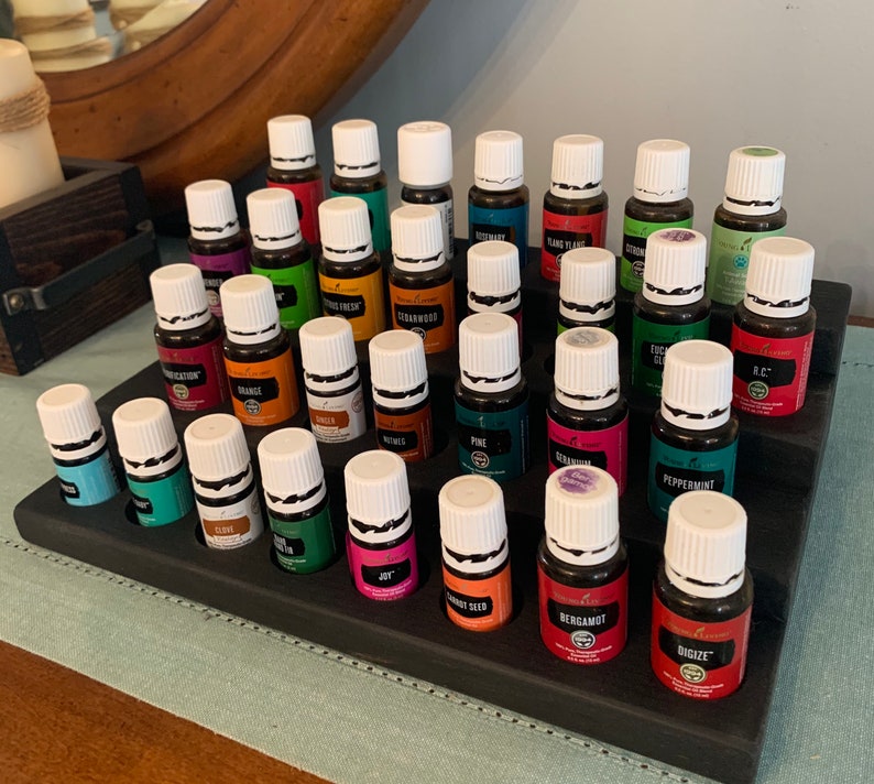 May include: A tiered, dark-stained wooden essential oil display stand holding numerous small glass bottles with white caps. The bottles are filled with various colored liquids and labeled with different essential oil names.