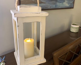 May include: A white wooden lantern with a rope handle and a lit candle inside. The lantern has a distressed finish and a rectangular shape. The candle is a cream color and emits a warm glow. The lantern is a decorative home accent.