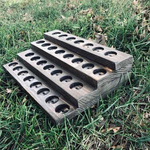 May include: A tiered wooden essential oil display stand with multiple circular openings. The stand is stained a dark gray color and is designed to hold small bottles. The stand is placed on a bed of green grass.