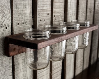 May include: A rustic, dark-stained wooden shelf holds five clear glass mason jars. The shelf is mounted on a weathered, light-colored wooden wall. The jars are empty and ready for use.