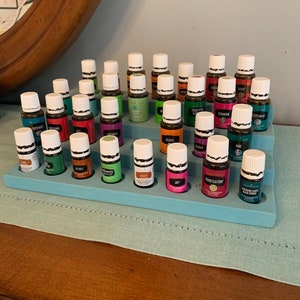 May include: A tiered, light blue wooden display shelf holding numerous small essential oil bottles. The bottles are various colors with white caps and labels. The shelf is on a wooden surface, with a mirror in the background.