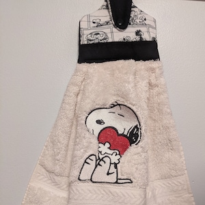 Puede incluir: Una toalla de cocina blanca con una imagen de dibujos animados en blanco y negro de Snoopy sosteniendo un corazón rojo. La toalla tiene un lazo de colgar negro con un patrón de dibujos animados en blanco y negro.