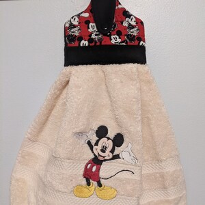 Puede incluir: Una toalla de cocina beige con un diseño de Mickey Mouse bordado. La toalla tiene un lazo de colgar negro y rojo con un estampado de Mickey Mouse.