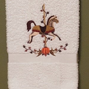 Puede incluir: Toalla de mano blanca con un diseño bordado de un caballo de carrusel con una calabaza y hojas de otoño.