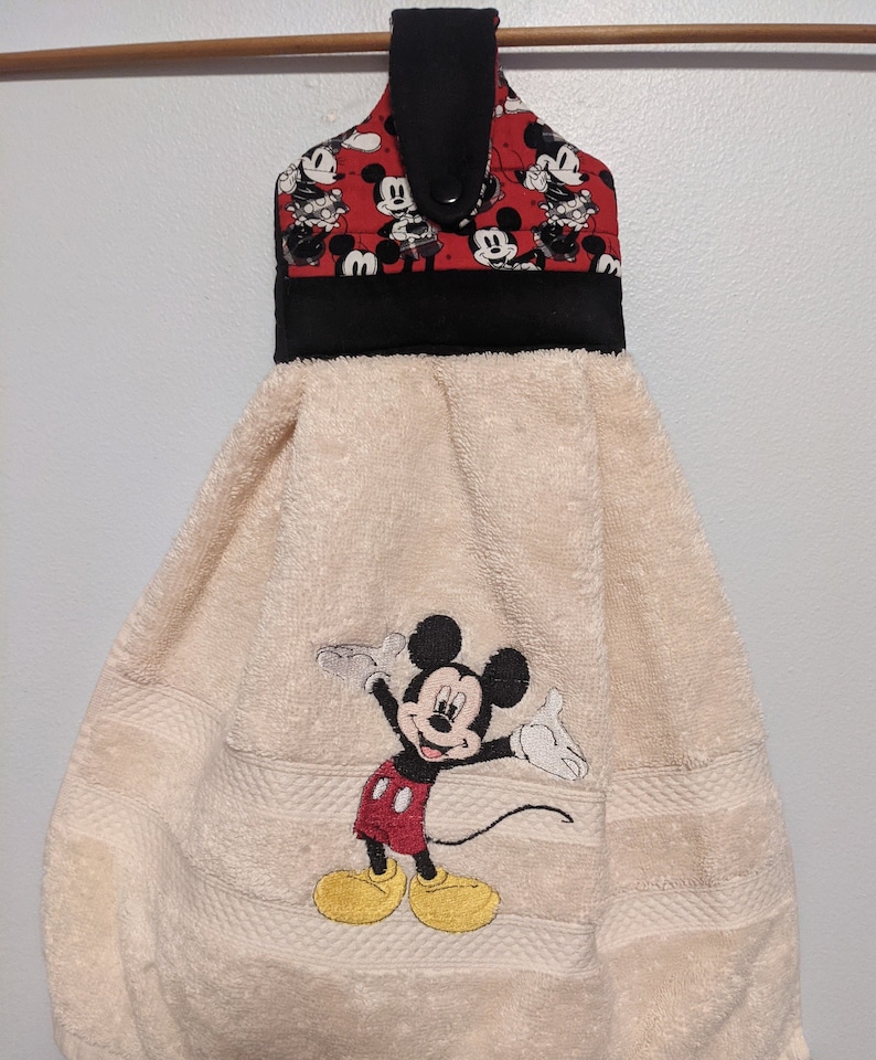 May include: A beige kitchen towel with a Mickey Mouse design embroidered on it. The towel has a black and red hanging loop with a Mickey Mouse print.