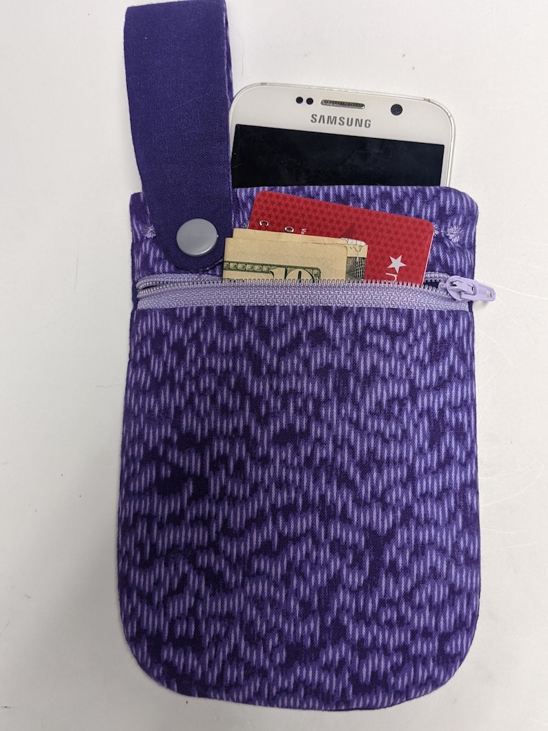 Puede incluir: Bolsa de color morado con cremallera que contiene un tel&eacute;fono, una tarjeta de cr&eacute;dito y dinero en efectivo. La bolsa tiene una correa morada con un bot&oacute;n plateado.