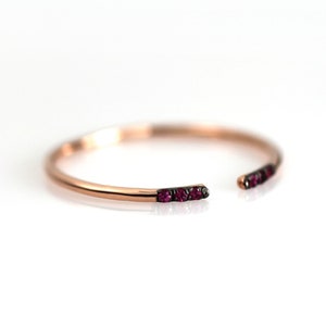 以下が含まれることがあります： 各端に小さな濃い赤色の宝石がセットされた、ローズゴールドのオープンカフブレスレット。 ブレスレットはシンプルでエレガントなデザインで、白い背景に表示されています。