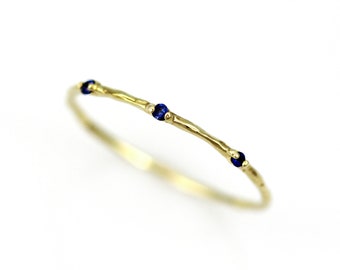 May include: A delicate gold ring with a textured band and three small, round, dark blue gemstones evenly spaced along the top. The ring is set against a bright white background.