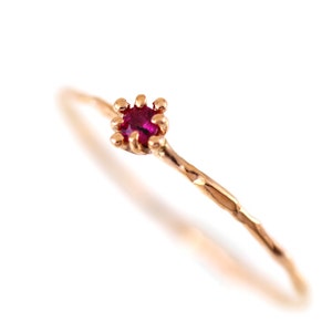 May include: A delicate gold ring with a small, vibrant red gemstone at the center. The ring's band has a textured, twig-like design. The gemstone is set in a crown-like setting, adding to its elegant appearance.