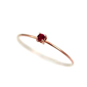 Könnte beinhalten: Ein zarter roségoldener Ring mit einem kleinen, runden, leuchtend roten Edelstein. Der Ring hat ein dünnes Band und ein schlichtes, elegantes Design. Der Edelstein ist in einer Vier-Zacken-Fassung gefasst.