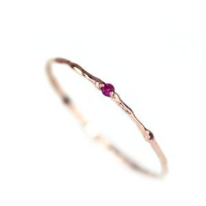 Könnte beinhalten: Ein zarter Ring aus Roségold mit einem strukturierten Band und einem kleinen, runden, leuchtend rosa Edelstein. Der Ring ist vor einem sauberen weißen Hintergrund platziert, der sein minimalistisches Design und seine elegante Schlichtheit hervorhebt. Schmuck.