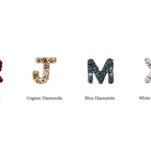 May include: Four letter-shaped jewelry pieces: an "R" with rubies, a "J" with cognac diamonds, an "M" with blue diamonds, and an "X" with white diamonds. Each letter is set against a white background, with the gemstone type labeled below each letter.