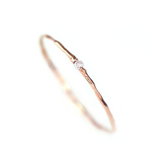 Könnte beinhalten: Ein zarter roségoldener Ring mit einem strukturierten Band und einem einzelnen, kleinen, klaren Edelstein. Der Ring ist vor einem hellweißen Hintergrund platziert, was seine schlichte Eleganz und sein minimalistisches Design hervorhebt.