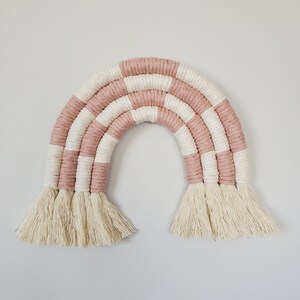May include: A pink and white striped macrame rainbow wall hanging with fringe tassels.