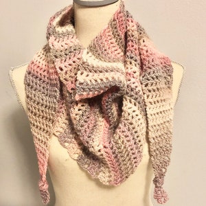 May include: A crocheted shawl in shades of pink, beige, and gray, with a delicate lace pattern. The shawl is draped over a mannequin and has tassels on the ends. The word "Carnation" is written below the shawl.
