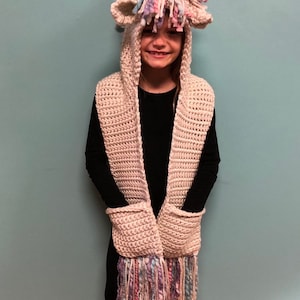 May include: A white crocheted unicorn hooded scarf with a pink, blue, and white yarn mane and horn. The scarf has pockets and a fringe.