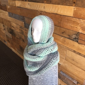 May include: A handmade, hooded scarf in shades of gray and mint green. The scarf is crocheted and draped over a mannequin head. The background is a wooden wall.