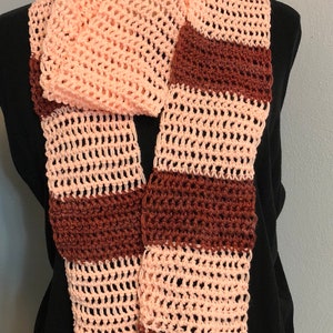 May include: A handmade crochet scarf in a light pink and brown striped pattern. The scarf has a fringed edge and is displayed on a mannequin. The open weave design adds a delicate touch to this cozy accessory.