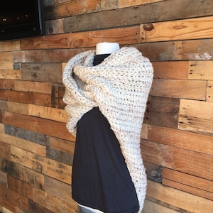 May include: A beige crocheted shawl with a textured pattern. The shawl is draped over a mannequin wearing a black shirt.