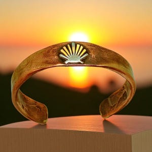 May include: A gold-toned cuff bracelet with a silver scallop shell design. The open-ended bracelet is displayed against a blurred background of a sunset. The bracelet is resting on a wooden surface.