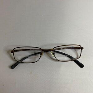May include: Brown rectangular eyeglasses with thin metal frames and black temple tips. The lenses feature a visible bifocal line. The inner frame is printed with 52 19-140 BROWN and the brand name Bulova Titanium Essex on the right lens.
