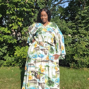 May include: A woman wearing a two-piece outfit with a tropical print. The top has wide sleeves and a matching belt. The skirt is long and pleated. The print features palm trees, flowers, and other tropical imagery in shades of blue, white, yellow, and green.