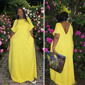 May include: A long, flowing yellow dress with short sleeves and pockets. The dress has a V-shaped back and is worn with a patterned tote bag. The background features a garden with pink flowers.