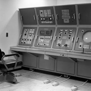 May include: A black and white photo of a control panel with a variety of buttons, knobs, and switches. A black swivel chair is in the foreground.