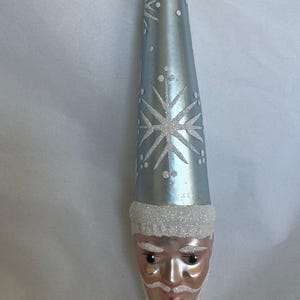May include: A blue glass Christmas ornament shaped like a Santa Claus head with a white snowflake design and a white glitter beard.