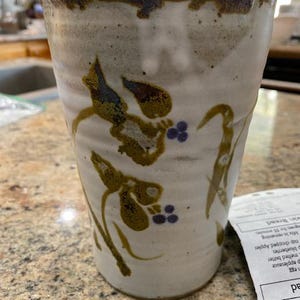 May include: A white ceramic mug with a brown rim and a floral design in shades of brown, green, and purple. The mug has a textured surface and a slightly flared lip.