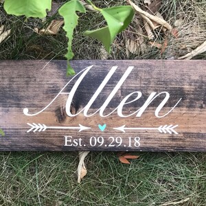May include: A rustic wooden sign with the name "Allen" written in white cursive script.  Two white arrows point towards a small turquoise heart with the text "Est. 09.29.18" below.