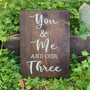 May include: A dark brown wooden sign with white text that reads "You & Me And Our Three".