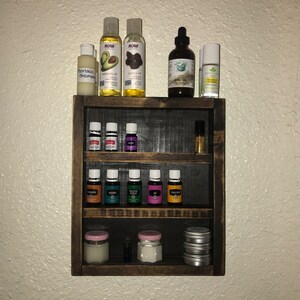 May include: A dark brown, three-tiered wooden shelf displaying various essential oils, lotions, and jars. The top shelf holds bottles of avocado oil, jojoba oil, and emu oil. The middle and bottom shelves contain small glass bottles and jars with pink lids.