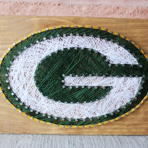 May include: Green Bay Packers logo string art on a wooden background. The logo is made of white and green string wrapped around nails. The logo is surrounded by a yellow border.